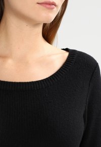 Černý žebrovaný svetr s širokým výstřihem do V, jemnou pleteninou a dlouhými rukávy, předvedený na části horní části těla modelky.