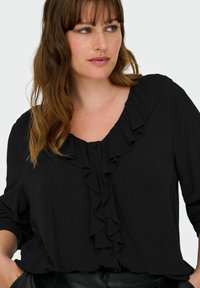 Melna blūze ar volānveida apkakli un pogu aizdari. Izgatavota no viegla auduma, tai ir garas piedurknes un brīvs siluets.