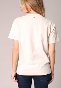 Camiseta de manga corta beige hecha de algodón, con un cuello redondo y un sutil detalle de logo en la parte superior de la espalda.