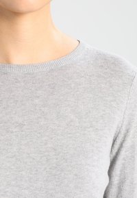 Pull gris clair en maille avec un col rond, présentant une texture douce et un design simple, sans motifs ni éléments métalliques visibles.