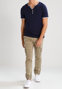 Marinblå kortärmad henley-tröja med tre vita knappar, kombinerad med beige cargobyxor och vita sneakers, med elastiska cuffs.