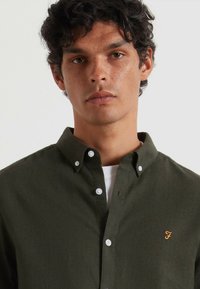 Ciemnozielona koszula na guziki, wykonana z gładkiego materiału. Posiada klasyczny kołnierz i białe guziki, a także małe haftowane logo w kolorze złotym.