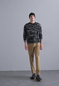 Schwarzer Pullover mit strukturiertem Schriftmuster, beige Hose und schwarzen Schuhen. Person steht vor einem schlichten grauen Hintergrund.