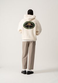 Crèmekleurige hoodie met een afbeelding van een boom en tekst op de achterkant. Gecombineerd met beige broek en zwarte schoenen, gezien van achteren.