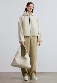 Μπουφάν από κρεμ δέρμα shearling με μεγάλες τσέπες, συνδυασμένο με μπεζ παντελόνι φαρδύ και τσάντα χρώματος κρεμ. Τα παπούτσια είναι λευκά, επίπεδα και απλά.