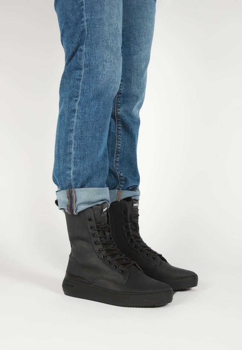 Personne portant un jean bleu retroussé et des bottines montantes noires à lacets, debout sur une surface blanche.