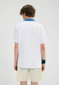 Wei�ßes Poloshirt mit kurzen Ärmeln und einem einfarbigen blauen Kragen, ausgestattet mit einem entspannten Schnitt und glatter Textur. Getragen mit hellgelben Shorts.