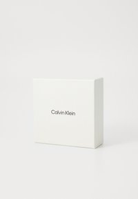 Biely obdĺžnikový box s nápisom "Calvin Klein" vytlačeným čiernym písmom na prednej strane, umiestnený na jednolatom bielom pozadí.