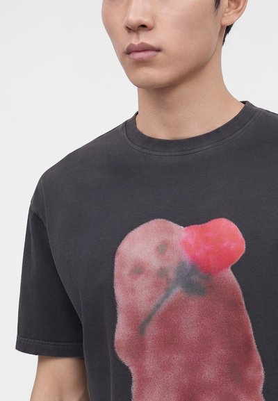 Νεαρός άνδρας φοράει μαύρο T-shirt με θολό κόκκινο γραφικό τριαντάφυλλο μπροστά, σε απλό φωτεινό φόντο.