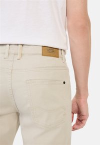 Beige Baumwollhosen verfügen über ein Leder-Logo-Patch, hintere Taschen mit subtilen Nähten und ein tailliertes Design.