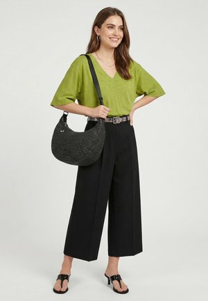 Donna sorride indossando una maglietta verde con scollo a V, pantaloni neri a gamba larga, sandali neri con tacco e una borsa a tracolla nera intrecciata.