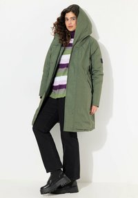 Abrigo impermeable largo de color verde oliva con capucha, combinado con un suéter a rayas multicolor y pantalones negros, con zapatos negros gruesos.