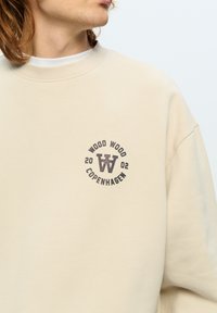 Beige sweatshirt met geribbelde ronde hals, met een circulaire grafiek in donkerbruin en de tekst "WOOD WOOD COPENHAGEN 2002" samen met het logo.