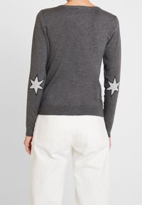 Person som bär en mörkgrå långärmad tröja med vita stjärnpatchar på armbågarna och vita högmidjade byxor, sedd bakifrån.