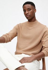 Suéter de punto beige con patrón texturizado, cuello de canalé y mangas acampanadas, combinado con pantalones vaqueros blancos. La modelo está sentada en una silla de madera.