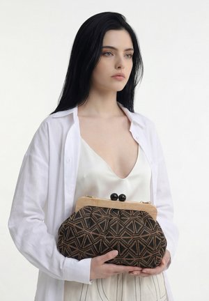 Giovane donna con lunghi capelli neri che indossa un top di seta bianco e una camicia tiene una pochette nera e marrone con motivo geometrico.