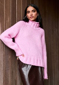 Rózsaszín vastag kötött pulóver magas bordázott gallérral és széles ujjakkal, barna műbőr nadrággal párosítva. Texturált, otthonos megjelenés.