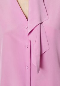 Blusa di raso rosa con scollatura a V, dotata di colletto legato, texture morbida e tre bottoni decorativi sul davanti.