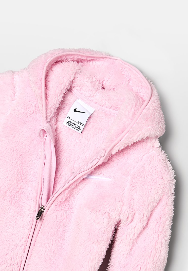 Sudadera con cremallera de felpa rosa, con una textura suave, que presenta una cremallera frontal y un pequeño logo blanco en el lado izquierdo. Etiqueta interior visible.