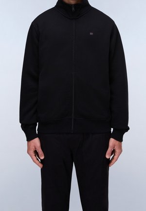 Mann trägt eine schwarze Jacke mit Reißverschluss, gerippten Bündchen und Saum, steht vor einem schlichten hellen Hintergrund mit entspannt hängenden Armen an den Seiten.