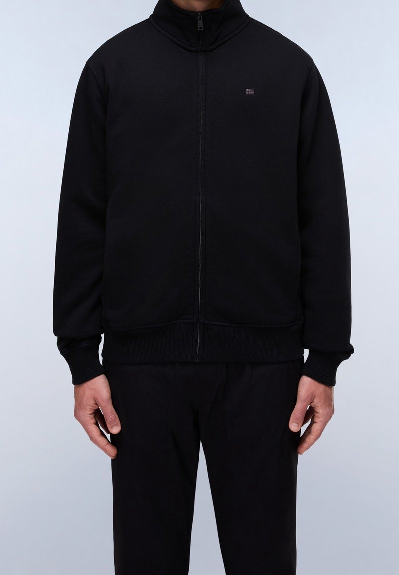 Homme portant une veste noire zippée avec des poignets et un ourlet côtelés, debout contre un fond clair uni, les mains détendues le long du corps.