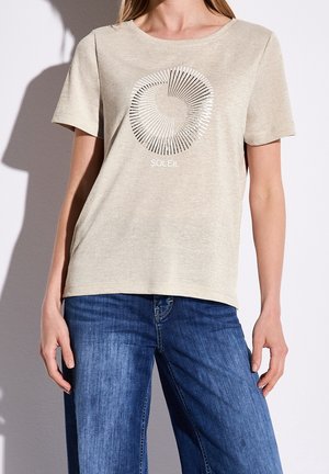 Lysebeige T-shirt med korte ærmer og rund hals, med grafisk solstråle i sølv. Kombineret med blå jeans.