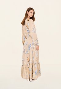 Blumenkleid mit langen Ärmeln in Beige, verziert mit blauen und orangefarbenen Blumen, mit gestuftem Rüschensaum und glattem Stoff. Modell steht und zeigt die Rückansicht.