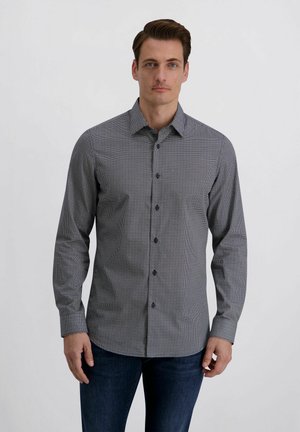 Mann trägt eng anliegendes, langärmeliges Hemd mit kleinem schwarz-weißem geometrischem Muster und dunkelblaue Jeans, steht vor neutralem Hintergrund.