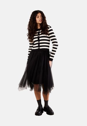 Cardigan rayé noir et blanc avec des boutons, associé à une jupe en tulle noire. Des chaussures noires chunky complètent le look.
