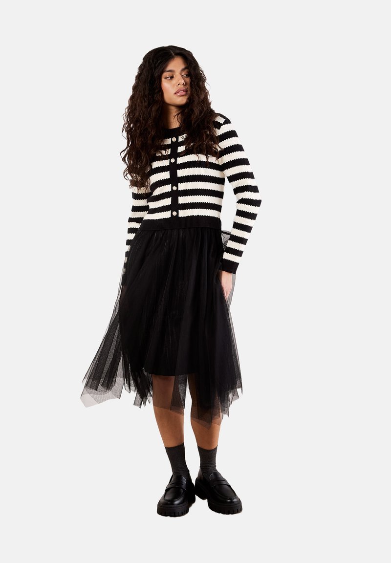 Cardigan a righe bianco e nero con bottoni, abbinato a una gonna di tulle nera. Scarpe nere massicce completano il look.