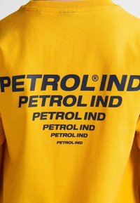 Tejido amarillo con el texto repetido "PETROL IND" en negro, disminuyendo de tamaño de arriba hacia abajo en la parte posterior de una prenda.