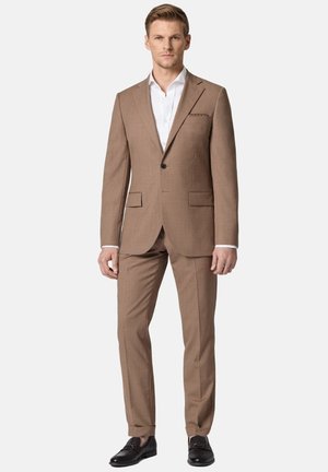 Mężczyzna ubrany w dopasowany jasnobrązowy garnitur, białą koszulę i czarne mokasyny, stojący na jasnym, jednolitym tle.