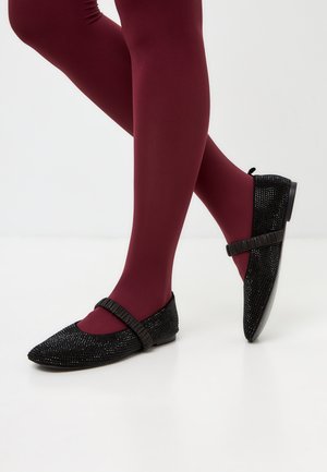 Zapatos planos negros con textura de lentejuelas, punta afilada y una correa elástica; llevados con medias burdeos sobre un fondo claro.