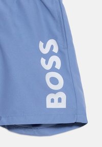 Ανοιχτό μπλε σορτς με λευκή επιγραφή "BOSS" τυπωμένη κάθετα κατά μήκος της πλαϊν ής ραφής και ορατή πλαϊνή τσέπη.