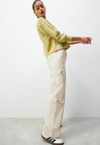 Pantaloni cargo bej cu multiple buzunare, asortați cu un pulover ușor verde. Ținuta are o croială lejeră și culori neutre.