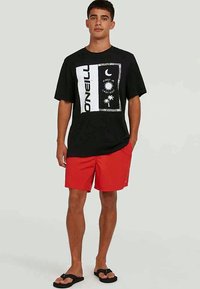 T-shirt gráfica preta com impressão branca, calções vermelhos e chinelos pretos; apresenta motivos de palmeiras e sol.
