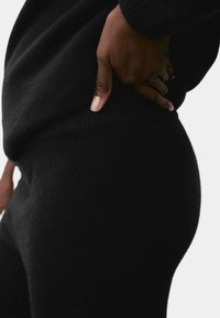 Prenda de punto suave y negra con ajuste ceñido, que cuenta con puños acanalados y una superficie texturizada. Mano colocada en la cadera, enfatizando la caída de la tela.