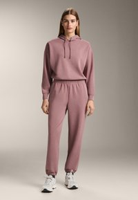 Rózsaszín pulóver és hozzáillő joggingnadrág puha pamut keverékből. Kapucni húzózsinórral, elasztikus derékpánt és mandzsetták. Sportos tornacipővel.