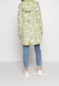 Grön regnjacka med vitt bladmönster, huva, dragsko i midjan och ventil bak. Matchad med ljusblå jeans och vita sneakers.