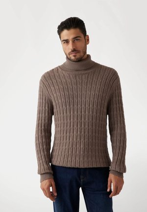 Brauner gerippter Strickpullover mit einem hohen Kragen und strukturiertem Muster; verfügt über lange Ärmel und eine lockere Passform, kombiniert mit blauen Jeans.