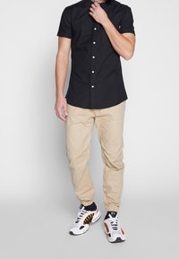 Homme portant une chemise à manches courtes noire avec boutons, un pantalon de jogging beige et des baskets blanches, noires et rouges, se tenant devant un fond uni.