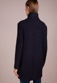 Abrigo de lana azul marino oscuro con cuello alto, tejido texturizado, hombros caídos y un corte de longitud media, con la espalda recta y sin cierres visibles.