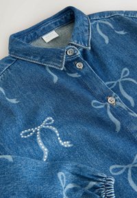 Jeanshemd in Blau, mit einem Button-Down-Kragen und hellgrauen Schleifenmustern. Zwei graue Knöpfe sind in der Nähe des Halsausschnitts sichtbar. Weicher Stoff.