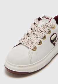 Baskets en cuir blanc avec des lacets texturés rouges et blancs, œillets dorés, et un accent de logo noir et blanc. Bout rond, semelle rembourrée.