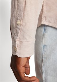 Donkerhuidige hand die ligt naast een lichtroze shirt met knopen en lichtblauwe denimjeans tegen een effen achtergrond.