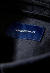 Etichetta in tessuto blu navy con la scritta "Lindbergh" in carattere serif bianco, cucita su un capo scuro testurizzato, con bordi chiari e cuciture ordinate.