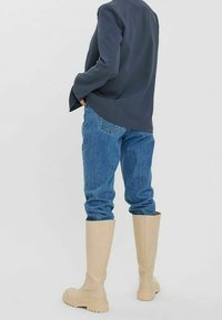 Beige knähöga stövlar med en slät textur, i kombination med ljusblå denimjeans och en mörkblå kavaj, sedd från baksidan.