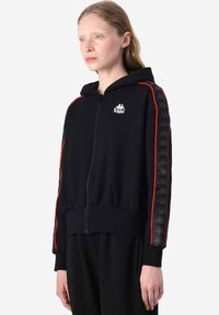 Felpa nera con zip e cappuccio, con strisce laterali rosse e logo Kappa sul petto. Presenta un design nero texturizzato lungo le maniche.