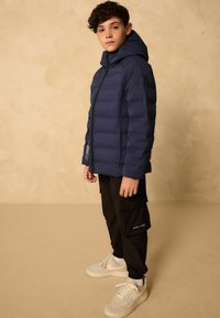 Garçon portant une doudoune bleu marine avec capuche, un pantalon cargo noir et des baskets blanches, se tenant devant un fond beige.