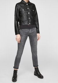 Chaqueta de cuero negra con cuello con solapa, dos bolsillos en el pecho y herrajes plateados. Combinada con jeans de mezclilla gris y botas negras con cordones.